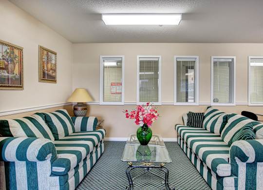 A cozy community lounge at **Quail Ridge** featuring two bold **green-and-white striped sofas** facing each other, a glass coffee table with floral decor, and soft overhead lighting. The space also includes framed wall art, a side table with a lamp, and interior windows that keep the room feeling open and connected.