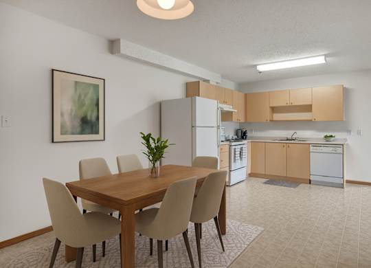 A bright dining area here at Quail Ridge with a wooden table, four upholstered chairs, and simple wall art for a clean, welcoming feel. A spacious kitchen sits just beyond with light wood cabinets, white appliances, and a neutral tile floor.