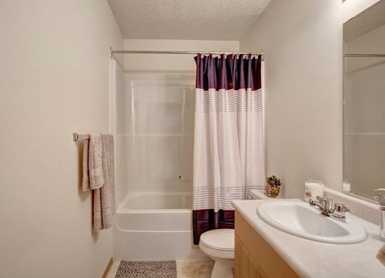A clean bathroom here at Quail Ridge with a bright bathtub-shower combo, a modern striped shower curtain, and soft neutral walls. A simple vanity with a white countertop and sink sits beneath a large mirror, creating a fresh and functional space.