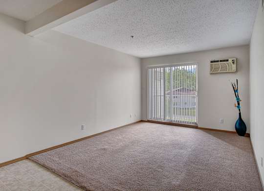A spacious, empty living area here at Quail Ridge with soft beige carpet and clean white walls for a bright, open feel. A large sliding glass door with vertical blinds brings in natural light, and a built-in wall AC unit adds everyday comfort.