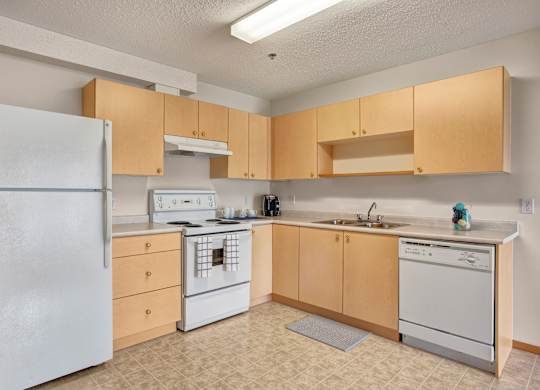 A bright, welcoming kitchen here at Quail Ridge with light wood cabinets, neutral countertops, and a classic white refrigerator and stove. A built-in dishwasher and stainless steel sink add everyday convenience, creating a clean and functional layout.