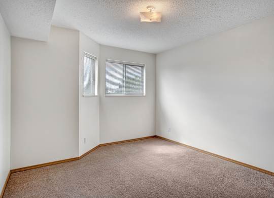 A bright, empty bedroom here at Quail Ridge with soft carpet, light walls, and natural light coming through wide windows with white blinds. A cozy corner nook adds extra space for a reading chair or small desk, creating a simple and inviting feel.