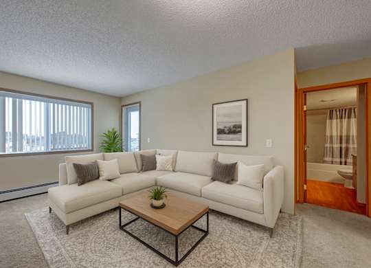 A bright, modern living room here at Sonora featuring a spacious neutral sectional sofa with accent pillows, a wooden coffee table on a soft area rug, large windows with vertical blinds bringing in natural light, and a clean, open layout connecting comfortably to the adjacent hallway and bathroom.