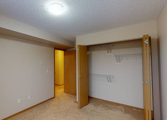 A spacious carpeted bedroom here at Sonora featuring neutral walls, a wide recessed ceiling detail, and an open double-door closet with built-in shelving and hanging space, complemented by warm wood trim, soft overhead lighting, and a layout that allows easy access to the hallway while offering flexible furniture placement.