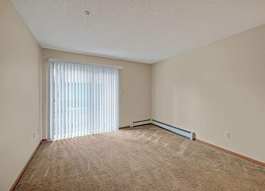 A bright carpeted living room here at Sonora featuring neutral walls, a large sliding glass door with vertical blinds that opens to a private balcony, baseboard heating along the wall, and an open rectangular layout that offers ample space for seating, media furniture, and everyday living.