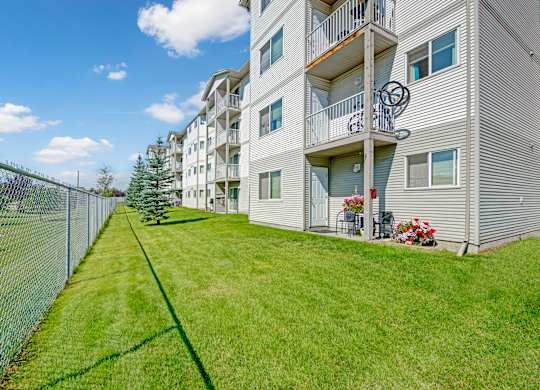 A landscaped exterior view here at Sonora showcasing a well-maintained green lawn alongside a multi-story apartment building with light-colored siding, private balconies on each level, patio seating with potted flowers, mature trees, and a fenced perimeter under a bright blue sky.