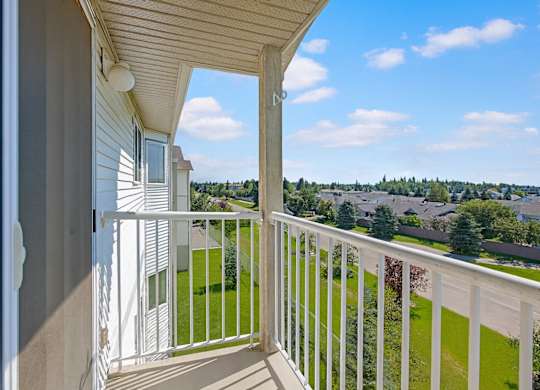 A private outdoor balcony here at Sonora featuring white railing, durable concrete flooring, and an elevated view overlooking lush green landscaping, mature trees, and a peaceful residential neighborhood under bright blue skies, offering a relaxing space to enjoy fresh air and scenic surroundings.