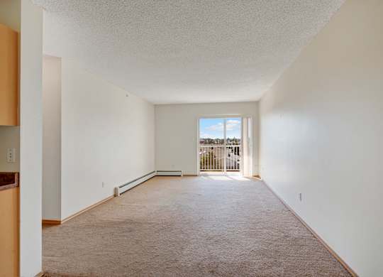 A bright and open living room here at Sonora featuring soft neutral carpeting, light cream walls, and a long rectangular layout that leads to a sliding glass door with vertical blinds opening to a private balcony, with ample natural light, a baseboard heater, and clean finishes creating a flexible and inviting space.
