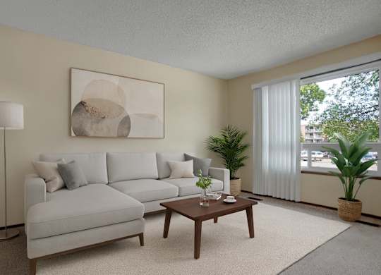 A bright modern living room here at Ventana featuring a light gray sectional sofa with neutral accent pillows, a wooden coffee table styled with minimal décor, a soft area rug, abstract wall art above the sofa, a tall floor lamp, and potted green plants near a large window with vertical blinds letting in natural light.