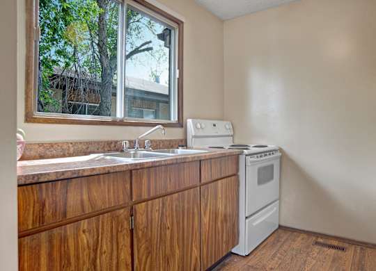 A bright kitchen here at Ventana featuring wood-style lower cabinetry, light laminate countertops, a double-basin sink beneath a large window with leafy views, white electric range and refrigerator, and wood-look flooring that adds warmth to the functional layout.