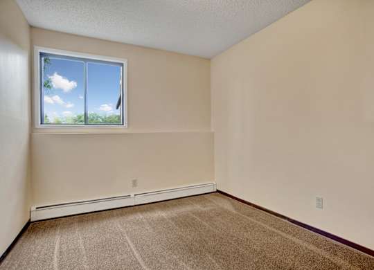 A cozy bedroom here at Ventana featuring soft neutral walls, plush carpet flooring, a baseboard heater along the wall, and a large window that brings in natural light and leafy outdoor views, creating a simple and comfortable space for rest.