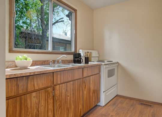 A bright functional kitchen here at Ventana featuring warm wood cabinetry, a laminate countertop with a double sink beneath a large window, a white electric range and refrigerator, vinyl plank flooring, and simple countertop accents, with natural light highlighting the clean layout and practical workspace.