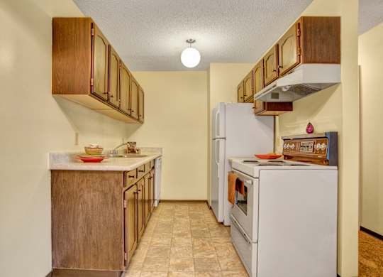 A warm compact kitchen here at Ventana showcasing rich wood cabinetry, a laminate countertop with integrated sink, white electric range and refrigerator, vinyl tile flooring, and simple staged accents, with overhead lighting and a streamlined galley layout that maximizes functionality and storage.