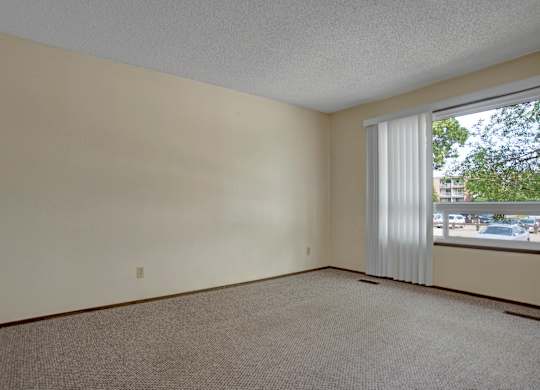 A spacious bedroom here at Ventana featuring neutral-toned walls, soft carpet flooring, a large window with vertical blinds that allow natural light, simple baseboard trim, and an open layout that offers flexibility for furniture placement and a bright, comfortable living environment.