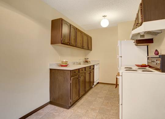 A warm galley kitchen here at Ventana featuring wood-style upper and lower cabinetry, light laminate countertops, white appliances including a refrigerator, range, and dishwasher, durable tile flooring, and soft overhead lighting that creates a functional and inviting cooking space.