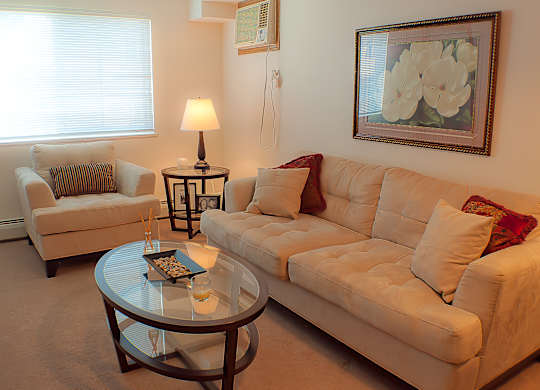 A cozy living room here at Wellington Manor with a cream sectional sofa, a matching armchair, and soft carpet flooring. A round glass coffee table and a warm side table lamp create a comfortable space for relaxing or entertaining. A large window with blinds brings in natural light, and a floral wall art piece adds a welcoming touch.