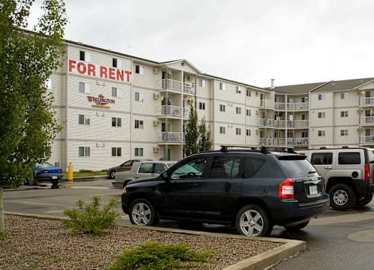 A wide exterior view here at Wellington Manor featuring a multi-story apartment community with private balconies and a large resident parking lot. A well-kept driveway and landscaped greenery create a welcoming first impression. A clear “For Rent” sign on the building makes it easy to spot the community from the street.