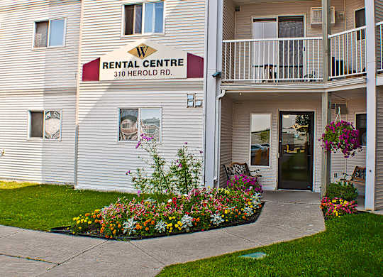 A welcoming rental office entrance here at Wellington Manor with a clearly marked Rental Centre sign and a covered walkway. A colorful landscaped flower bed and hanging baskets add vibrant curb appeal along the front path. A seating area near the door creates a friendly spot for guests and residents.