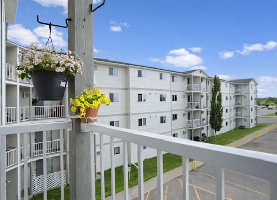 A sunny balcony view here at Wellington Manor overlooking the community buildings, green landscaping, and the parking area below. A white railing and hanging flower baskets add a charming touch to the outdoor space. A bright blue sky creates a peaceful and open atmosphere.