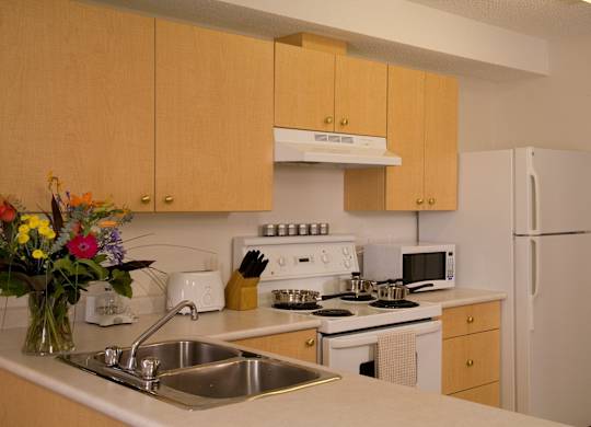A bright kitchen here at Wellington Manor with light wood cabinets, clean white countertops, and a double stainless steel sink. A white stove with a vent hood, a microwave, and a full-size refrigerator provide a practical cooking setup. A wide breakfast bar counter adds extra prep space and a convenient spot for casual dining.