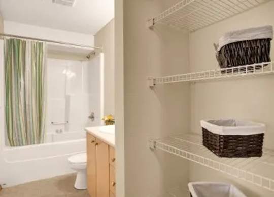 A clean bathroom here at Wellington Manor with a bathtub-shower combo, a simple striped shower curtain, and a white toilet. A light wood vanity with a built-in sink adds a warm and practical touch. A spacious open shelving area with wire racks provides convenient storage for towels and everyday essentials.