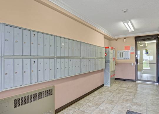 A clean community mailroom here at Wellington Manor with a long wall of secure resident mailboxes and bright overhead lighting. A tiled floor and neutral wall tones create a neat and well-kept space. A convenient entry door and parcel area provide easy access for everyday mail pickup.