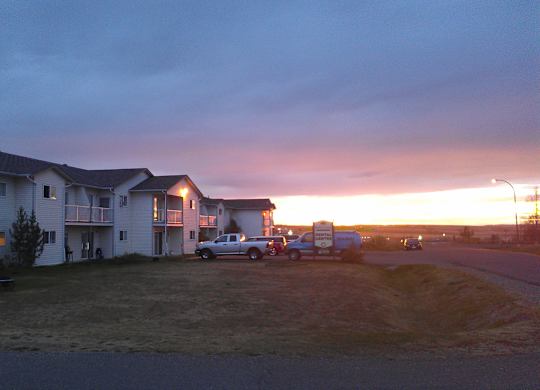 A wide community view here at Willowview Terrace featuring a row of light-sided apartment buildings with balconies and open green space in the foreground. Parked vehicles and a rental office sign sit along the road, while the soft sunset sky adds a warm, peaceful backdrop.