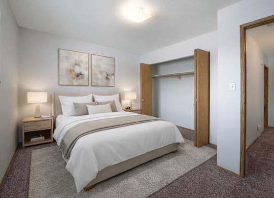 A bright, cozy bedroom here at Willowview Terrace featuring a neatly made bed with white bedding and soft neutral accent pillows. Matching nightstands with simple lamps sit on each side, while two modern wall art pieces add a clean touch above the headboard. An open closet and plush carpet complete the simple, comfortable layout.