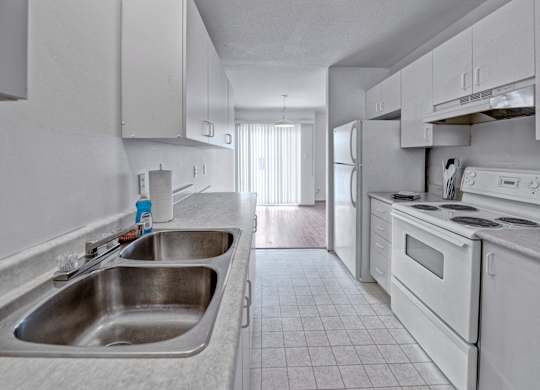 A bright galley-style kitchen here at Willowview Terrace with white cabinets, light countertops, and a double stainless steel sink. White appliances including a stove, range hood, and refrigerator line the space, with durable tile flooring that leads into the dining area with a sliding glass door for natural light.