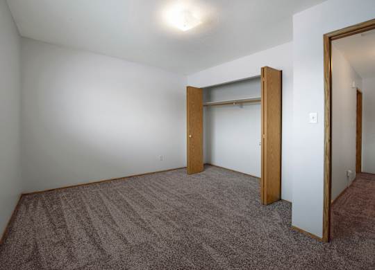 A simple, bright bedroom here at Willowview Terrace with soft carpeting, white walls, and a ceiling light for even lighting. An open closet with wood sliding doors and a hanging rod provides practical storage, with a hallway entrance off to the side for an easy layout.