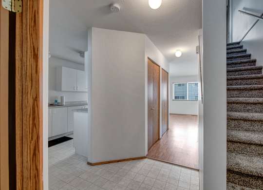 A bright entry hallway here at Willowview Terrace with light tile flooring, warm wood closet doors, and a clear walkway leading into the main living area. Carpeted stairs with a simple handrail add easy access to the upper level, while the nearby kitchen cabinetry keeps the layout practical and open.