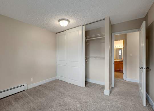 A cozy bedroom here at Wyndham Crossing with soft neutral walls, plush carpet flooring, and a ceiling light for a bright, simple space. A wide sliding-door closet with built-in shelving and hanging storage sits near the hallway leading to the bathroom vanity area.
