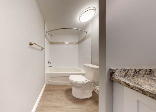 A simple bathroom here at Wyndham Crossing featuring a white tub and shower combination, a curved shower rod, and bright overhead lighting. A white toilet sits beside the tub, while wood-look flooring and a granite-style countertop add a clean, modern finish.