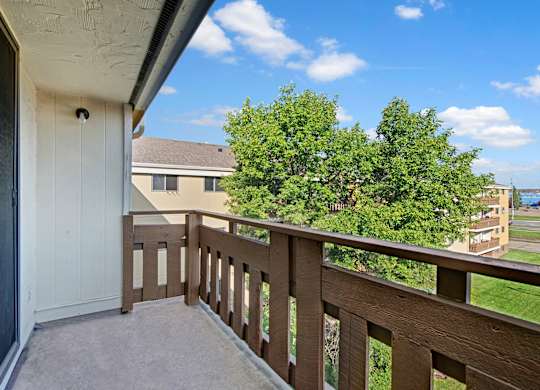 A private balcony view here at Wyndham Crossing with a concrete patio floor and a sturdy brown railing overlooking green trees and nearby buildings. The bright blue sky with soft clouds adds a fresh, open feel to the outdoor space.