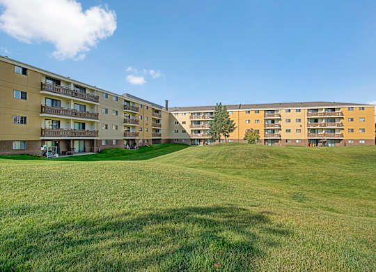 A wide exterior view here at Wyndham Crossing showcasing two multi-story apartment buildings with private balconies and a large open green lawn in between. A few mature trees and clear blue skies create a calm, open setting around the community.