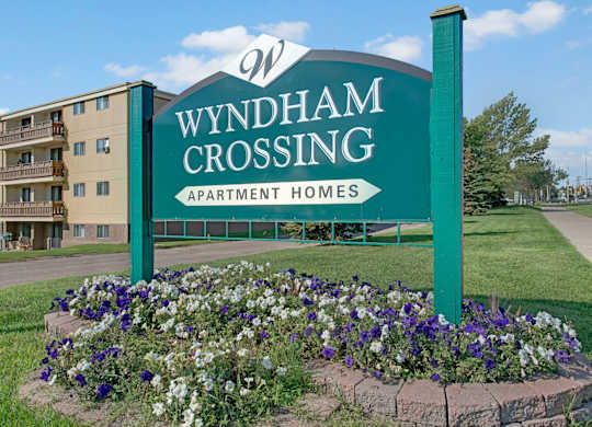 A welcoming community entrance sign here at Wyndham Crossing featuring a large green “Wyndham Crossing Apartment Homes” display surrounded by a colorful flower bed. A multi-story apartment building and neatly kept green lawn sit in the background under a bright blue sky.