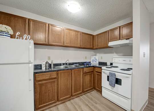 A cozy kitchen here at Wyndham Crossing with warm wood cabinets, dark countertops, and white appliances including a stove and refrigerator. A simple sink area with small countertop decor and wood-style flooring completes the clean and functional layout.