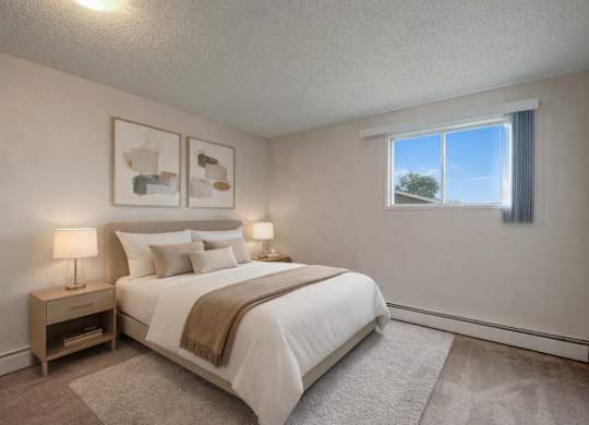 A bright, relaxing bedroom here at Wyndham Crossing with a cozy bed dressed in crisp white bedding, neutral accent pillows, and a soft beige throw blanket. Two matching nightstands with modern lamps sit on either side, while simple wall art and a large window bring in natural light above the plush carpet.