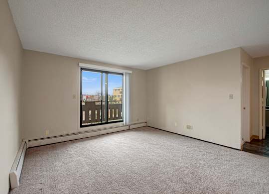 A bright living room here at Wyndham Crossing with soft carpet flooring, light neutral walls, and a wide sliding glass door that opens to a private balcony. An open layout with baseboard heating and an adjacent hallway entry creates a comfortable space for relaxing or entertaining.