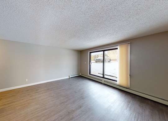 A bright open living room here at Wyndham Crossing with wood-style flooring, soft neutral walls, and a large sliding window that brings in natural light. The long layout offers plenty of space for a comfortable seating area, with simple baseboard heating along the window wall.