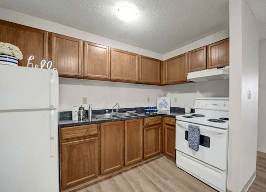A cozy kitchen here at Wyndham Crossing with warm wood cabinets, dark countertops, and white appliances including a stove and refrigerator. A simple sink area with small countertop decor and wood-style flooring completes the clean and functional layout.