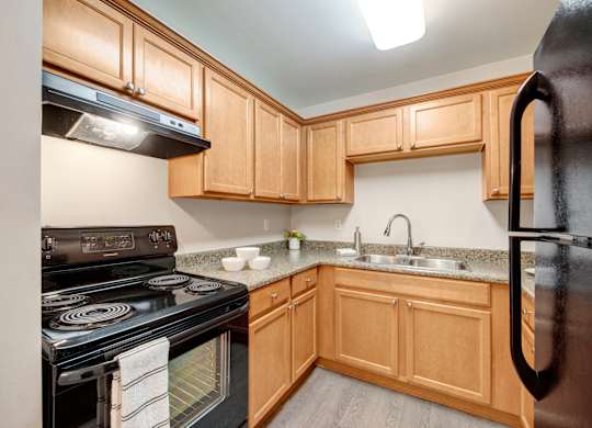 A bright kitchen here at Wyndham Crossing with warm wood cabinetry, speckled countertops, and a double-basin sink with a modern faucet. Black appliances and a smooth wood-style floor add a clean, updated look with plenty of storage and prep space.
