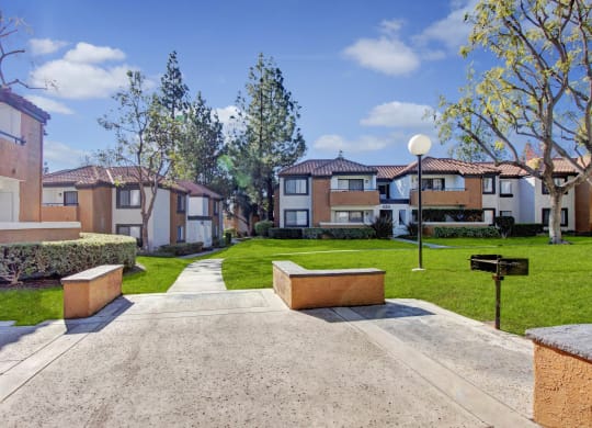 Sunlit Castleparks Apartment complex with terracotta roofs and white walls surrounds a grassy courtyard. A walkway, trees, and a barbecue grill create a serene atmosphere.