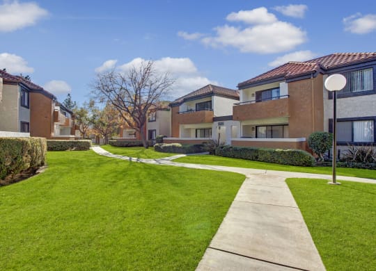Pathway through a well-manicured lawn flanked by modern beige and brown Castlepark Apartment Homes buildings, under a bright blue sky with scattered clouds. Peaceful suburban setting.