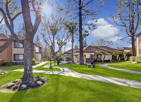 A sunlit outdoor scene of at our Castlepark Apartment Homes complex with well-kept grass, trees, and colorful flowerbeds. A sign reads "Leasing Office," conveying a welcoming tone.