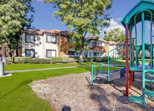 Playground here at Castlepark Apartment Homes with green jungle gym in foreground on mulch, surrounded by lush grass and trees, with beige two-story residential buildings in background.