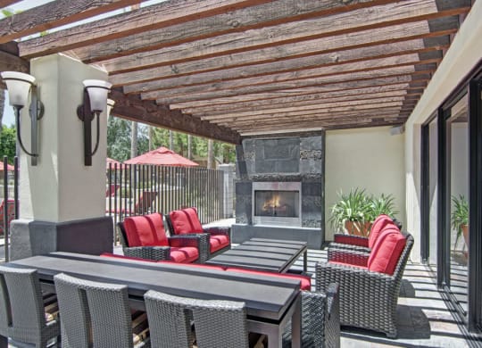 Outdoor patio here at Natura Villas Apartments with a wooden pergola, featuring red-cushioned wicker chairs, a black table, a modern stone fireplace, and potted plants. Sunny and inviting.