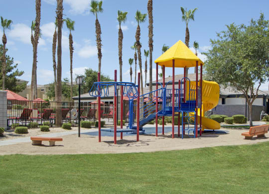 Colorful playground here at Natura Villas Apartments with a yellow slide and red-blue climbing structures on sand, surrounded by palm trees and greenery, under a clear blue sky.