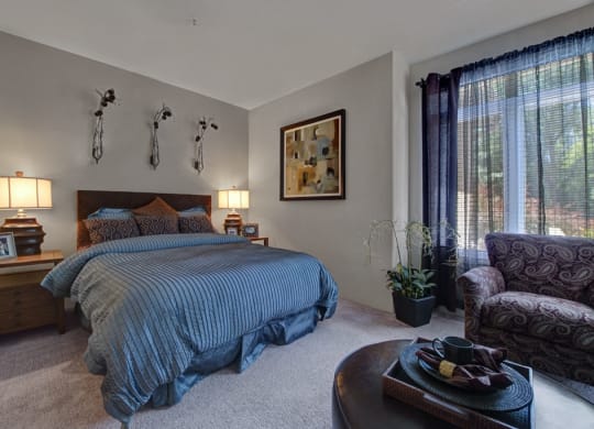 Cozy bedroom here at Cheyenne Creek Apartment Homes with a blue-striped bed, two wooden nightstands with lamps, abstract wall art, large window with curtains, and patterned armchair. Warm ambience.