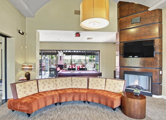 Modern lounge area here at Natura Villas Apartments with a curved orange sofa and square-patterned pillows, set before a fireplace and TV. Large window reveals an outdoor patio.